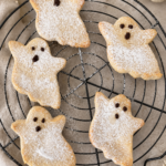 Homemade ghost-shaped shortcrust cookies dusted with icing sugar and decorated with chocolate faces, arranged on a cooling rack.